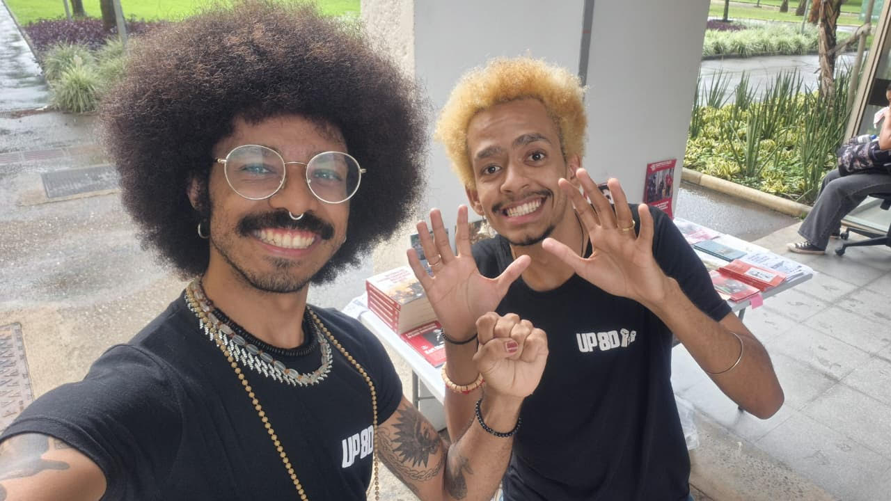 Dois jovens negros sorridentes posam para uma selfie em frente a uma mesa montada com livros e jornais. O jovem à esquerda tem cabelo black power, usa óculos redondos e colares. O jovem à direita tem o cabelo loiro e faz um gesto com as mãos. Ambos vestem camisetas pretas com a inscrição "UP"