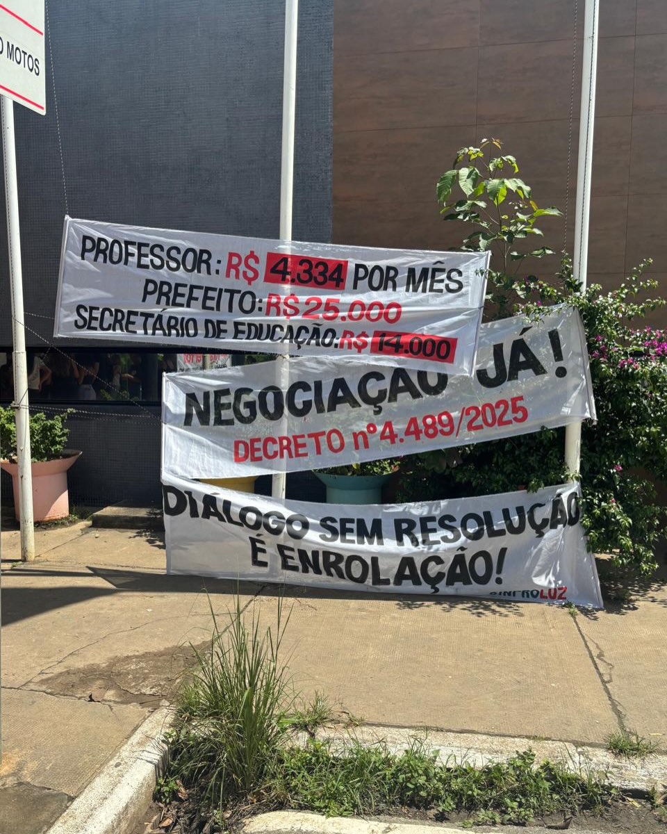 Fotografia vertical de três faixas brancas estendidas entre mastros em uma calçada. A primeira faixa no topo detalha a desigualdade salarial em letras pretas e vermelhas: "Professor: R$4.334 por mês, Prefeito: R$25.000; Secretário de Educação: R$ 14.000". A faixa do meio diz: "Negociação Já! Decreto nº 4.489/2025". A faixa inferior traz a frase: "Diálogo sem resolução é enrolação!". No canto inferior direito da última faixa, lê-se a assinatura do sindicato Sinproluz.