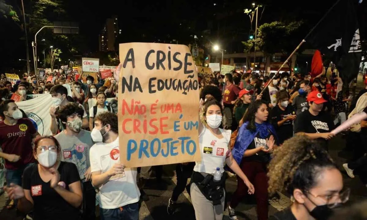 Manifestação noturna em defesa da educação pública. Em destaque, uma jovem segura um cartaz de papelão com os dizeres: "A crise na educação não é uma crise é um projeto". Ao fundo, manifestantes usam máscaras e carregam bandeiras de movimentos populares em uma via urbana iluminada.