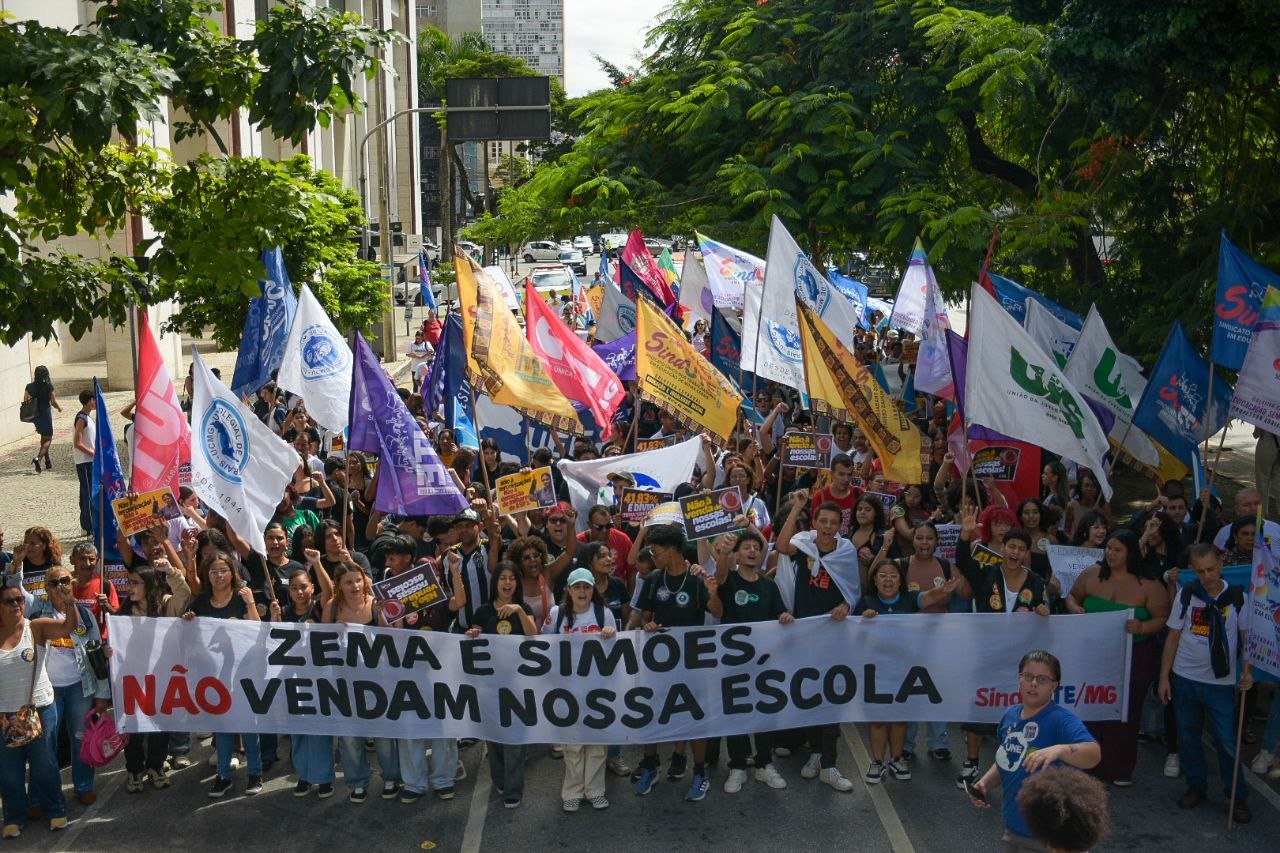 Fotografia de uma manifestação em defesa da educação pública em Minas Gerais contra o leilão de escolas promovido pelo governo Zema. Em primeiro plano, manifestantes seguram uma grande faixa branca com os dizeres "ZEMA E SIMÕES, NÃO VENDAM NOSSA ESCOLA" em letras pretas e vermelhas. Ao fundo, uma multidão agita diversas bandeiras coloridas de sindicatos e movimentos estudantis.