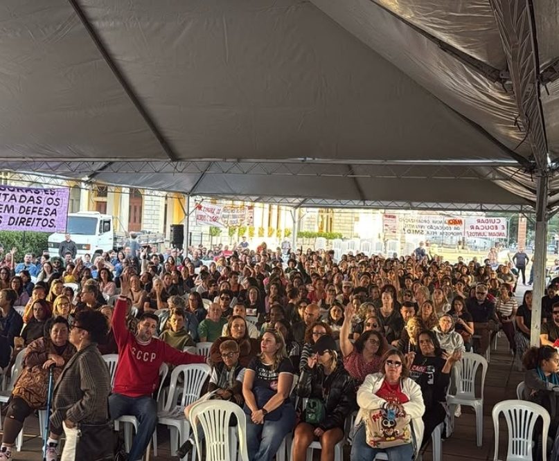 Fotografia de uma assembleia de trabalhadores em educação de Belo Horizonte. Sob uma grande tenda cinza, centenas de pessoas estão sentadas em cadeiras brancas de plástico voltadas para uma frente comum. Algumas pessoas estão com os braços levantados, sugerindo um momento de votação. Ao fundo, veem-se faixas de protesto penduradas e parte de um prédio histórico.